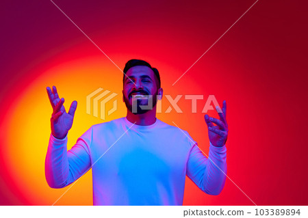 Portrait of handsome bearded man smiling, laughing, posing with positive mood against gradient studio background in neon light Portrait of handsome bearded man smiling, laughing, posing with positive mood against gradient studio background in neon light 103389894