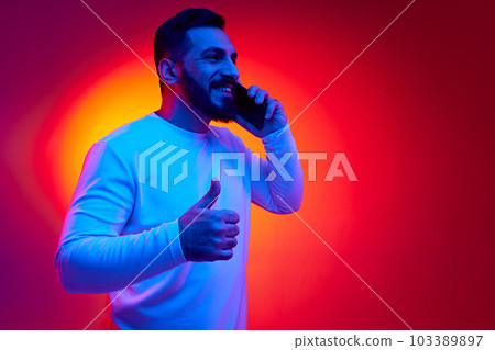 Portrait of handsome bearded man in casual clothes talking on mobile phone against gradient studio background in neon light. Approvement 103389897