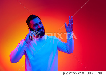 Happy talk. Portrait of handsome bearded man in casual clothes talking on mobile phone against gradient studio background in neon light 103389906