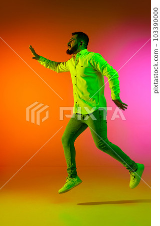 Full-length portrait of handsome bearded man in casual clothes jumping against gradient studio background in neon light Full-length portrait of handsome bearded man in casual clothes jumping against gradient studio background in neon light 103390000