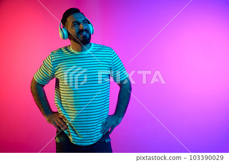 Portrait of bearded man in striped shirt listening to music in headphones against gradient pink blue studio background in neon light 103390029
