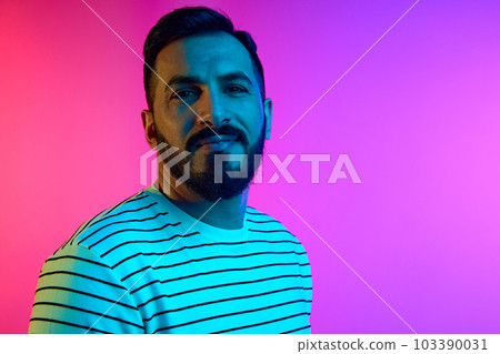 Portrait of handsome mature bearded man in striped shirt looking at camera with tender smile against gradient pink blue studio background in neon light 103390031