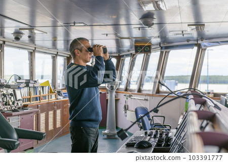 Deck officer with binoculars on navigational bridge. Seaman on board of vessel. Commercial shipping. Cargo ship. Deck officer with binoculars on navigational bridge. Seaman on board of vessel. Commercial shipping. Cargo ship. 103391777