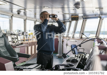 Deck officer with binoculars on navigational bridge. Seaman on board of vessel. Commercial shipping. Cargo ship. 103391782