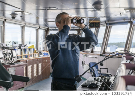 Deck officer with binoculars on navigational bridge. Seaman on board of vessel. Commercial shipping. Cargo ship. 103391784