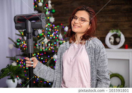 An attractive girl holds a vacuum cleaner in her hand against the background of a Christmas tree. 103391801
