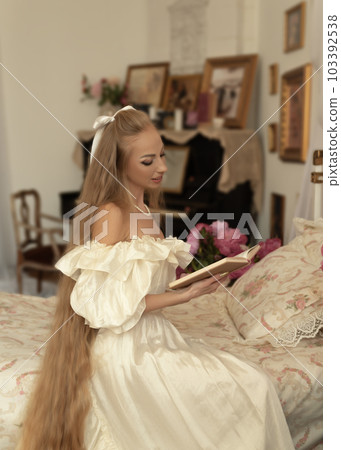 A beautiful young woman with thick long hair sits on a bed in a room with paintings and flowers and reads a book.19th century. Vintage. 103392538