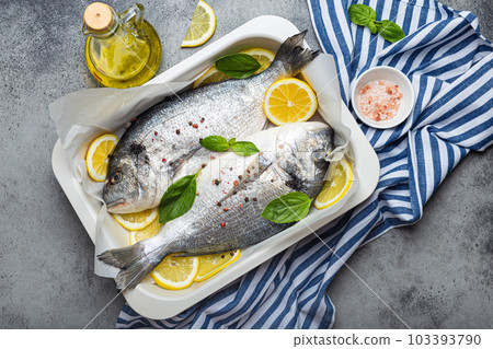 Raw fish dorado in casserole dish with ingredients lemon, fresh basil, bottle of olive oil on wooden cutting board with knife on rustic stone background top view, cooking healthy fish dorado Raw fish dorado in casserole dish with ingredients lemon, fresh basil, bottle of olive oil on wooden cutting board with knife on rustic stone background top view, cooking healthy fish dorado 103393790