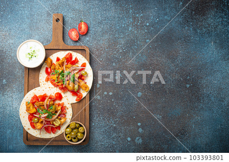 Cooking Traditional Greek Dish Gyros: Pita bread with vegetables, meat, herbs, olives on rustic wooden cutting board with Tzatziki sauce, olive oil top view on dark blue stone background, copy space. 103393801