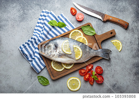 Raw uncooked fish dorado with ingredients lemon, fresh basil, cut cherry tomatoes on wooden cutting board with knife on rustic stone background top view, cooking healthy fish dorado Raw uncooked fish dorado with ingredients lemon, fresh basil, cut cherry tomatoes on wooden cutting board with knife on rustic stone background top view, cooking healthy fish dorado 103393808