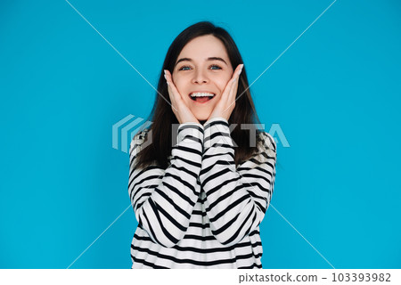 Expressive Portrait of a Surprised and Joyful Young Woman with Palms on Cheekbones, Isolated on Blue 103393982