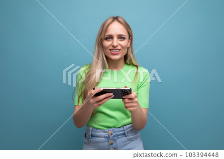 adorable blond girl in casual outfit plays a game on the phone on a blue background adorable blond girl in casual outfit plays a game on the phone on a blue background 103394448