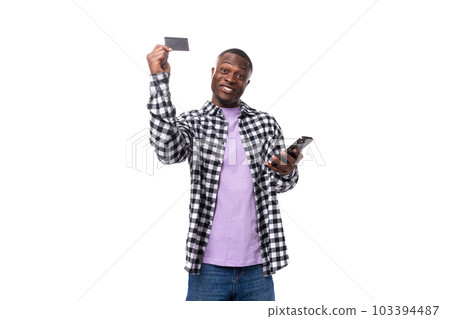positive smiling young african man with short haircut in plaid shirt holding card mockup and phone 103394487