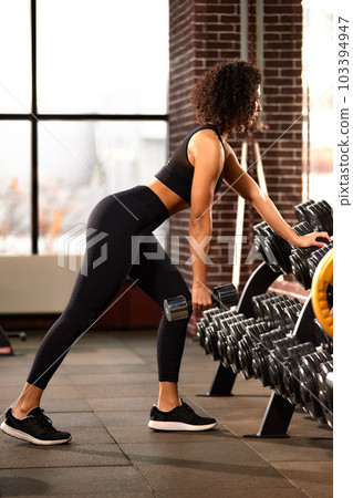 African American woman training with dumbbells in the hall in front of the mirror, against large windows, bright hall, positive view of the woman ternating the muscles of the hands 103394947