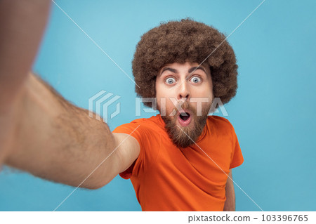 Portrait of surprised shocked man with Afro hairstyle wearing orange T-shirt taking selfie or broadcasting livestream, looking with wow expression, POV. Indoor studio shot isolated on blue background. Portrait of surprised shocked man with Afro hairstyle wearing orange T-shirt taking selfie or broadcasting livestream, looking with wow expression, POV. Indoor studio shot isolated on blue background. 103396765