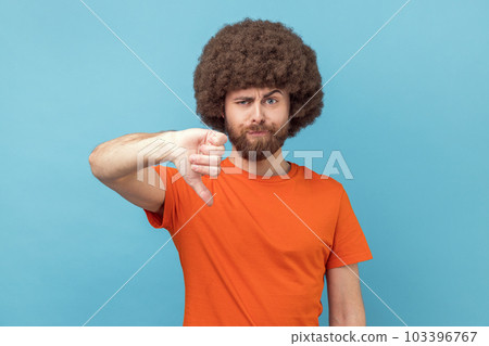 Portrait of displeased man with Afro hairstyle wearing orange T-shirt showing thumbs down dislike gesture, symbol of disagree, giving feedback. Indoor studio shot isolated on blue background. Portrait of displeased man with Afro hairstyle wearing orange T-shirt showing thumbs down dislike gesture, symbol of disagree, giving feedback. Indoor studio shot isolated on blue background. 103396767
