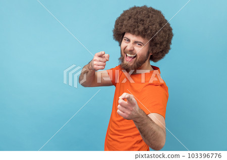 Portrait of man with Afro hairstyle wearing orange T-shirt pointing to camera, smiling, making happy choice, we need you concept. Indoor studio shot isolated on blue background. Portrait of man with Afro hairstyle wearing orange T-shirt pointing to camera, smiling, making happy choice, we need you concept. Indoor studio shot isolated on blue background. 103396776