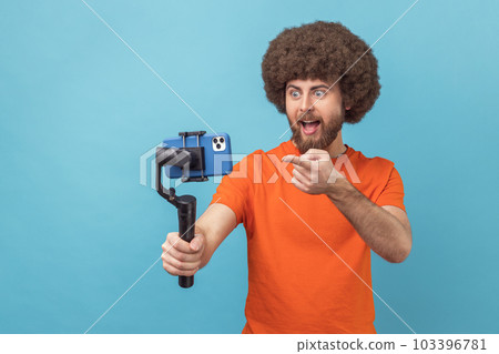 Portrait of astonished man with Afro hairstyle wearing orange T-shirt using stedicam and phone for livestream, being amazed, pointing at phone camera. Indoor studio shot isolated on blue background. 103396781