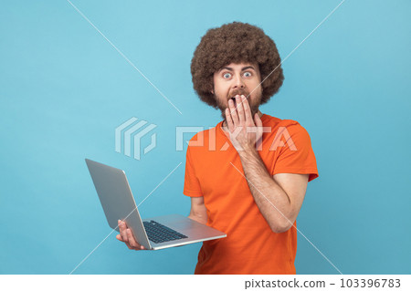Portrait of man with Afro hairstyle wearing orange T-shirt working on notebook, sees something astonishing, covering mouth with palm. Indoor studio shot isolated on blue background. 103396783