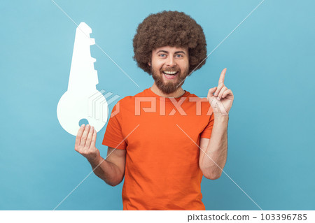 Portrait of smiling man with Afro hairstyle wearing orange T-shirt holding big paper key and pointing finger up, having idea for renting new flat. Indoor studio shot isolated on blue background. Portrait of smiling man with Afro hairstyle wearing orange T-shirt holding big paper key and pointing finger up, having idea for renting new flat. Indoor studio shot isolated on blue background. 103396785