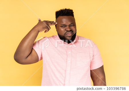 Kill me pleas. Portrait of desperate man wearing pink shirt pointing finger gun to head, shooting herself, making suicide gesture to stop depression. Indoor studio shot isolated on yellow background. 103396786