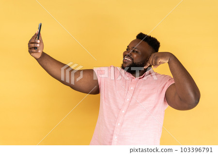 Portrait of handsome positive man wearing pink shirt taking selfie or having livestream, pointing finger down, subscribe his vlog. Indoor studio shot isolated on yellow background. Portrait of handsome positive man wearing pink shirt taking selfie or having livestream, pointing finger down, subscribe his vlog. Indoor studio shot isolated on yellow background. 103396791