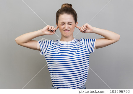 I dont want to hear this. Annoyed frustrated woman wearing striped T-shirt closing ears with fingers ignoring information, tired of irritating noise. Indoor studio shot isolated on gray background. 103396793