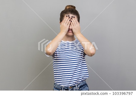 Don't want to look at this. Portrait of confused woman wearing striped T-shirt covering eyes with hand, feeling shamed and scared to watch. Indoor studio shot isolated on gray background. 103396797