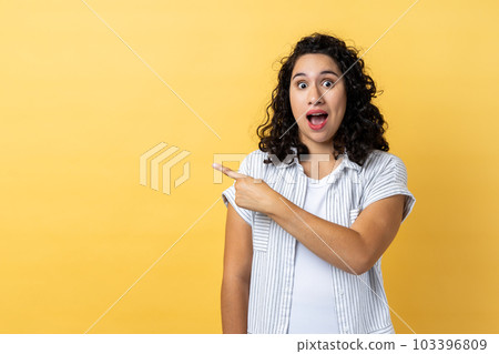 Portrait of amazed shocked beautiful woman with dark wavy hair standing pointing aside with finger, showing copy space for advertisement. Indoor studio shot isolated on yellow background. Portrait of amazed shocked beautiful woman with dark wavy hair standing pointing aside with finger, showing copy space for advertisement. Indoor studio shot isolated on yellow background. 103396809