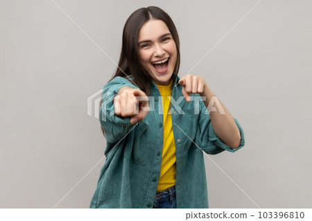 Hey you. Positive dark haired young woman pointing to camera, showing direction and choosing, indicating with index finger, wearing casual style jacket. Indoor studio shot isolated on gray background. Hey you. Positive dark haired young woman pointing to camera, showing direction and choosing, indicating with index finger, wearing casual style jacket. Indoor studio shot isolated on gray background. 103396810