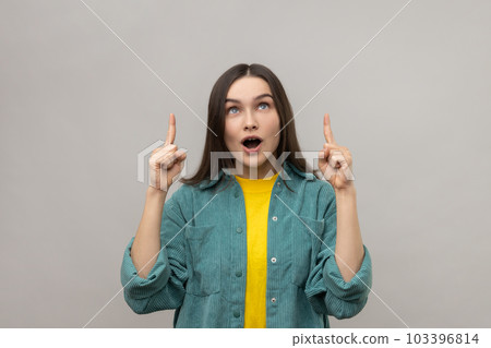 Portrait of surprised woman with open mouth pointing fingers up at empty copy space, place for advertisement, wearing casual style jacket. Indoor studio shot isolated on gray background. Portrait of surprised woman with open mouth pointing fingers up at empty copy space, place for advertisement, wearing casual style jacket. Indoor studio shot isolated on gray background. 103396814
