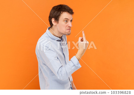 Side view portrait of angry rude man standing looking away with aggression, showing middle finger, fighting with someone, wearing light blue shirt. Indoor studio shot isolated on orange background. 103396828