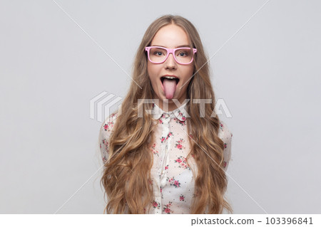 Portrait of childish carefree woman in glasses with long blond hair showing out tongue and closing eyes with naughty disobedient grimace, making face. Indoor studio shot isolated on gray background. 103396841