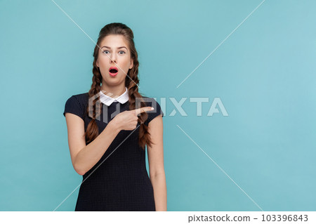 Portrait of shocked surprised woman points away on copy space, suggests follow this direction or click on link, wearing black dress. woman Indoor studio shot isolated on blue background. Portrait of shocked surprised woman points away on copy space, suggests follow this direction or click on link, wearing black dress. woman Indoor studio shot isolated on blue background. 103396843