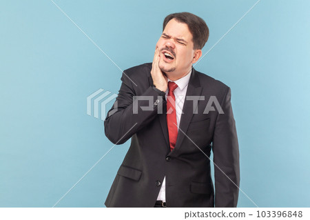 Portrait of man suffers painful toothache, has rotten tooth, needs to visit dentist, keeps eyes closed from pain, wearing black suit with red tie. Indoor studio shot isolated on light blue background. Portrait of man suffers painful toothache, has rotten tooth, needs to visit dentist, keeps eyes closed from pain, wearing black suit with red tie. Indoor studio shot isolated on light blue background. 103396848