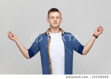 Portrait of calm meditative handsome teenager boy wearing blue shirt standing with closed eyes and yoga gesture pose, trying to calm down. Indoor studio shot isolated on gray background. Portrait of calm meditative handsome teenager boy wearing blue shirt standing with closed eyes and yoga gesture pose, trying to calm down. Indoor studio shot isolated on gray background. 103396849