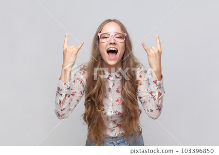 Rock n roll. Portrait of woman in glasses with wavy blond hair standing with rock sing, screaming yelling with excited facial expression. Indoor studio shot isolated on gray background. 103396850
