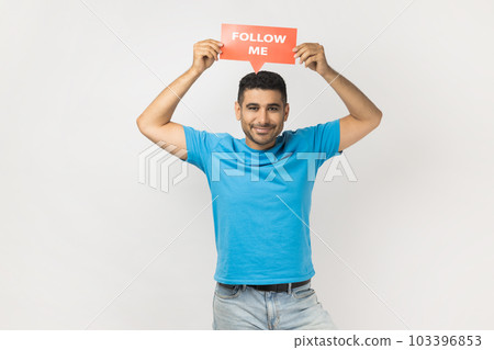 Portrait of joyful smiling handsome unshaven man wearing blue T- shirt standing holding card with follow me inscription above head. Indoor studio shot isolated on gray background. 103396853