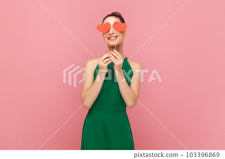 Portrait of charming unknown woman with bun hairstyle standing and covering her eyes with little red hearts on stick, smiling, wearing green dress. Indoor studio shot isolated on pink background. 103396869