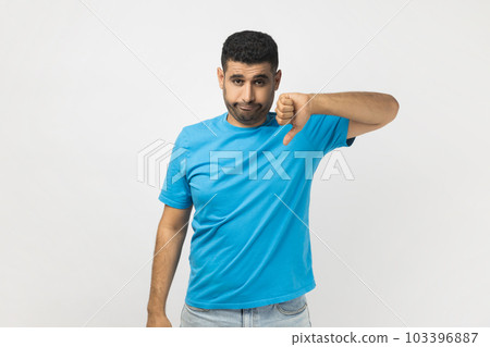 Portrait of unshaven man wearing blue T- shirt standing assess project, shows sign of dislike, looks with negative expression and disapproval. Indoor studio shot isolated on gray background. 103396887
