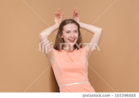 Portrait of funny smiling joyful woman with long hair standing with raised arms, showing horns, looking at camera, wearing elegant dress. Indoor studio shot isolated on brown background. Portrait of funny smiling joyful woman with long hair standing with raised arms, showing horns, looking at camera, wearing elegant dress. Indoor studio shot isolated on brown background. 103396888