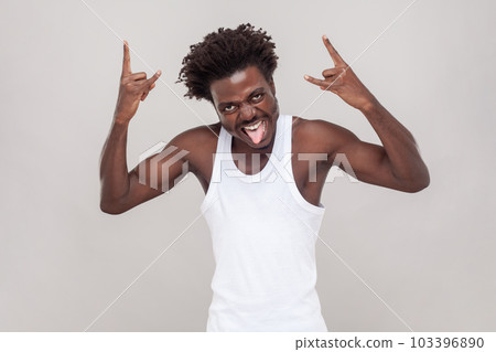 Man with Afro hairstyle shouting from happiness, has widely opened mouth, makes rock n roll gesture, says rock this party. wearing white T-shirt. Indoor studio shot isolated on gray background. Man with Afro hairstyle shouting from happiness, has widely opened mouth, makes rock n roll gesture, says rock this party. wearing white T-shirt. Indoor studio shot isolated on gray background. 103396890
