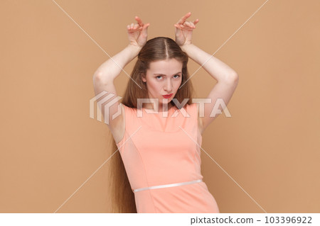 Portrait of funny positive woman with long hair standing with raised arms, showing horns, looking at camera, wearing elegant dress. Indoor studio shot isolated on brown background. 103396922