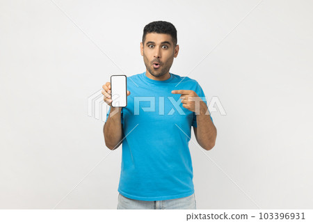 Portrait of shocked amazed unshaven man wearing blue T- shirt standing pointing at smart phone with blank white display, mockup for promotional text. Indoor studio shot isolated on gray background. Portrait of shocked amazed unshaven man wearing blue T- shirt standing pointing at smart phone with blank white display, mockup for promotional text. Indoor studio shot isolated on gray background. 103396931