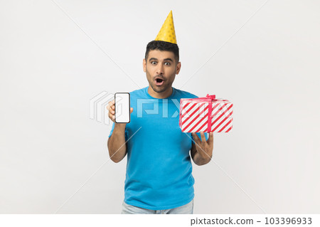 Shocked astonished surprised unshaven man wearing blue T- shirt and yellow party cone holding present box and showing cell phone with empty display. Indoor studio shot isolated on gray background. Shocked astonished surprised unshaven man wearing blue T- shirt and yellow party cone holding present box and showing cell phone with empty display. Indoor studio shot isolated on gray background. 103396933