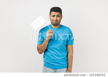 I give up. Portrait of unshaven man wearing blue T- shirt standing and holding white flag and looking at camera with sad face, crying. Indoor studio shot isolated on gray background. I give up. Portrait of unshaven man wearing blue T- shirt standing and holding white flag and looking at camera with sad face, crying. Indoor studio shot isolated on gray background. 103396935