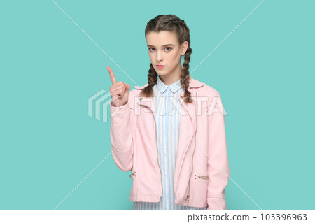 Portrait of serious bossy teenager girl with braids wearing pink jacket standing with raised finger, warning her classmates about danger. Indoor studio shot isolated on green background. 103396963