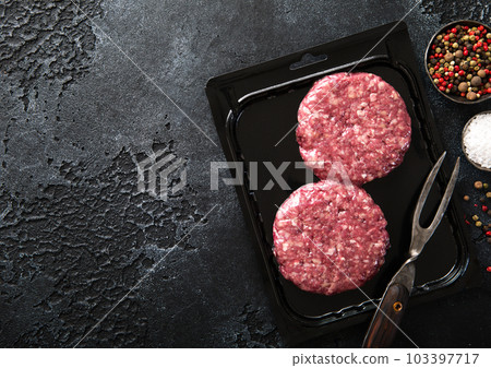 Raw organic beef burgers sealed in vacuum tray with fork and pepper on dark kitchen Raw organic beef burgers sealed in vacuum tray with fork and pepper on dark kitchen 103397717