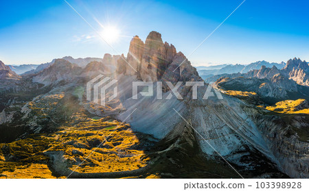 Landscape of rocky Three Peaks of Lavaredo at sunrise. Curved roads with cars on highland slopes around mountain under blue sky aerial view Landscape of rocky Three Peaks of Lavaredo at sunrise. Curved roads with cars on highland slopes around mountain under blue sky aerial view 103398928