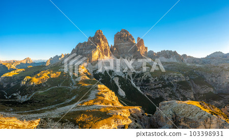 Rocky peaks of Tre Cime di Lavaredo at sunrise. Mountain landscape of wide hilly canyons and valley with serpentine roads under clear sky aerial view Rocky peaks of Tre Cime di Lavaredo at sunrise. Mountain landscape of wide hilly canyons and valley with serpentine roads under clear sky aerial view 103398931